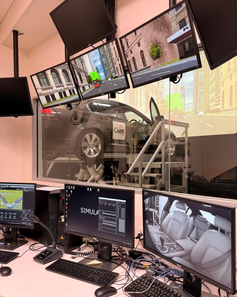 Driving simulator lab with a car on a motion platform, seen from the control room with many monitors.<br />
